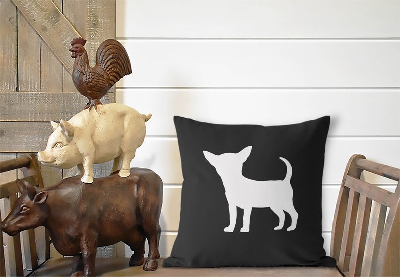 May include: A black throw pillow with a white silhouette of a chihuahua dog. The pillow is sitting on a wooden chair with a brown cow, pig, and rooster figurine stacked on top of each other.