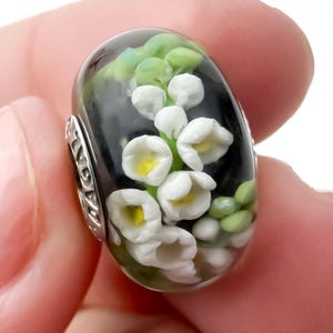 Peut inclure: Une perle de verre avec un fond noir, représentant des fleurs de muguet blanches avec des centres jaunes et des feuilles vertes. La perle a des accents métalliques argentés sur les côtés.
