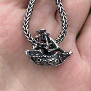 May include: A silver charm in the shape of a ship with a detailed design. The charm is attached to a silver chain.