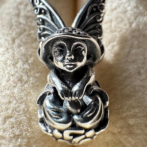 May include: A detailed silver pendant depicting a whimsical fairy figure. The fairy has large, ornate wings, a hat, and is holding a small object. The pendant is intricately designed with a focus on texture and detail, showcasing a fantasy theme.