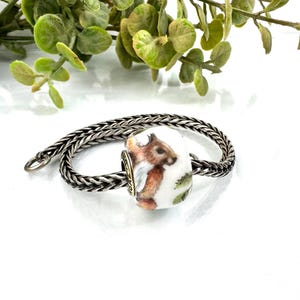 以下が含まれることがあります： 手描きのリスが描かれた白い磁器のビーズが付いたシルバーチャームブレスレット。ビーズには、ふさふさの尻尾と緑の葉を持つ茶色のリスが描かれています。