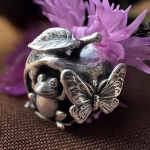 Könnte beinhalten: Eine silberfarbene Metallperle in Form eines modellierten Apfels mit einem Blatt oben, einem kleinen Frosch, der an der Seite hervorlugt, und einem detaillierten Schmetterling auf der Vorderseite. Das Stück hat ein metallisches Finish mit dunklen Vertiefungen.