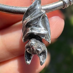 May include: A silver-toned metal charm shaped like a bat hanging upside down with its wings folded around its body. The charm features detailed textures on the wings and head, and it is attached to a smooth, rounded silver-toned metal bracelet band.