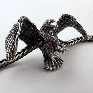 Könnte beinhalten: Eine detaillierte Silberadlerskulptur mit ausgebreiteten Flügeln, die auf einer geflochtenen Metallkette sitzt. Die Federn des Adlers sind aufwendig geschnitzt, und seine Krallen sind sichtbar. Die Skulptur ist vor einem schlichten weißen Hintergrund platziert.