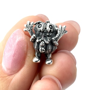 May include: A small, silver-colored charm in the shape of a pumpkin-headed figure. The charm has a textured surface, with details like a carved face and outstretched arms. The figure is standing on two small feet.