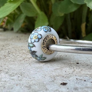 May include: A white bead with a floral design featuring blue flowers and green leaves. The bead has a gold-colored ring with the word "DAGAZ" on it. The bead is on a silver-colored metal bar.