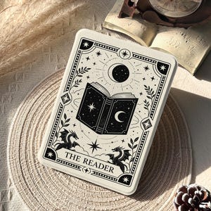 May include: A white rectangular tarot card featuring a black illustration of an open book, stars, a crescent moon, and dragons. The text "THE READER" is at the bottom. The card is on a beige woven placemat with a clock and open book in the background.