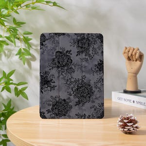 Può includere: Una custodia grigia per tablet con un motivo floreale nero. Il design presenta grandi fiori stilizzati e delicate viti su uno sfondo screziato. La custodia è rettangolare e sembra essere realizzata in un materiale liscio.