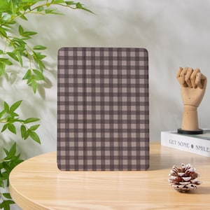 May include: A tablet case featuring a brown and beige gingham pattern. The case is rectangular with rounded edges, placed on a light brown wooden table. A wooden hand sculpture and a pinecone are also on the table.