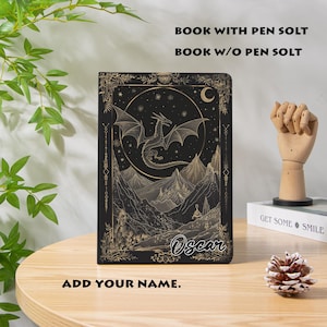 May include: Black book with a dragon and mountain design. The cover features a dragon with outstretched wings, a crescent moon, and the text "Oscar". The book has the text "BOOK WITH PEN SOLT" and "BOOK W/O PEN SOLT" and "ADD YOUR NAME."