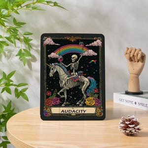 Può includere: Una stampa artistica rettangolare nera con uno scheletro che cavalca un unicorno scheletrico sotto un arcobaleno. Lo scheletro tiene in mano dei calici di vino. La stampa è incorniciata con rose e la parola "AUDACITY" è in basso.