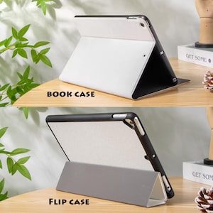 May include: Two tablet cases, one white and black, labeled "BOOK CASE," and the other gray and black, labeled "FLIP CASE." Both cases are shown open, displaying their interior and design. The cases are on a wooden surface with greenery in the background.