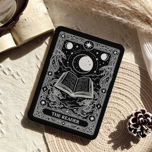 May include: A rectangular black card featuring a white line art illustration of an open book floating under a large moon and stars, surrounded by four dragons. The card is framed by an ornate border and includes the text THE READER at the bottom.
