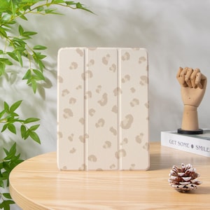 May include: A beige tablet case featuring a leopard print pattern. The case is closed, with a tri-fold design. The leopard print has light brown spots on a beige backdrop. The case rests on a wooden table.