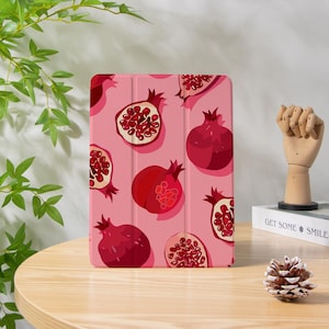 May include: A tablet case with a pink background featuring a pattern of red pomegranates. The pomegranates are whole and sliced, showcasing the seeds. The case is on a wooden table.