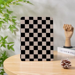 May include: A tablet case featuring a black and beige chequered design. The case is rectangular with rounded corners. It rests on a wooden table, alongside a wooden hand sculpture and a pinecone.