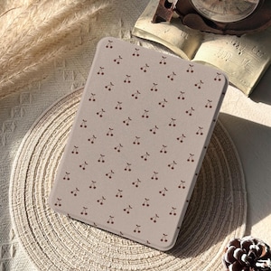 Puede incluir: Una funda de tableta beige claro con un patrón repetido de pequeñas cerezas marrones. La funda rectangular con esquinas redondeadas está colocada sobre un salvamanteles tejido. El logotipo de Glow Dwell Designs es visible en la esquina inferior izquierda.