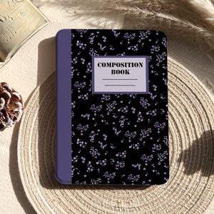 Op de afbeelding: Een compositieboek met een zwarte kaft versierd met een delicaat bloemenpatroon in paarse tinten. De rug is effen paars en de kaft heeft de tekst "COMPOSITION BOOK" in het wit.