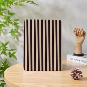 May include: A rectangular item featuring a black and beige vertical stripe design. It rests on a light brown wooden surface. A wooden hand sculpture and a pine cone are also visible.