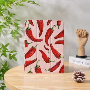 Può includere: Una custodia per tablet con un motivo ripetuto di peperoncini rossi con steli verdi su sfondo rosa chiaro. I peperoncini sono stilizzati con ombreggiatura dettagliata. La custodia è esposta su una superficie di legno.