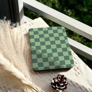 Könnte beinhalten: Ein quadratischer Untersetzer mit einem grün-dunkelgrünen Schachbrettmuster. Der Untersetzer liegt auf einer cremefarbenen, strukturierten Oberfläche. Ein Tannenzapfen und dekoratives Gras sind ebenfalls vorhanden.