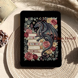 May include: A black rectangular e-reader cover featuring an embroidered illustration of a dragon perched on a stack of books, surrounded by red and yellow roses, green ivy, and small star and moon symbols. The text Glow Dwell Designs is visible.