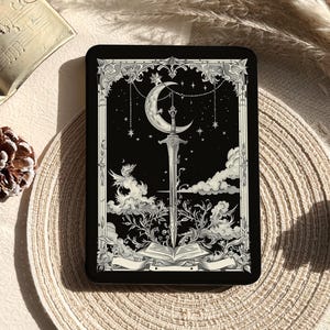 May include: A rectangular black tin featuring a detailed white illustration of a sword piercing an open book, set against a night sky with a crescent moon, stars, and clouds, all framed by an ornate border with decorative foliage and a blank banner.