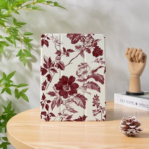 Può includere: Una custodia per tablet color crema con un motivo floreale bordeaux. Il design comprende grandi fiori, foglie e steli, creando un'estetica vintage. La custodia è esposta su un tavolo di legno con una pigna e un libro che dice "GET SOME SMILE".