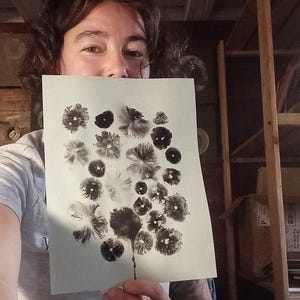 Puede incluir: Una persona sostiene un papel con un diseño floral en blanco y negro. La obra de arte presenta varias formas y patrones de flores, creando un tema botánico. El fondo incluye estanterías de madera y una camiseta gris.