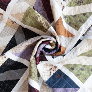 May include: A handcrafted quilt with a patchwork design. The quilt features a variety of fabrics in shades of brown, green, purple, and cream, arranged in geometric patterns. The quilt is folded, showcasing its intricate details and textures.