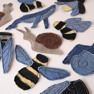May include: A collection of fabric patches featuring various animal shapes including snails made of brown corduroy, bees with black and yellow stripes, a blue denim whale, and blue denim insects with stitched wing details and frayed edges.