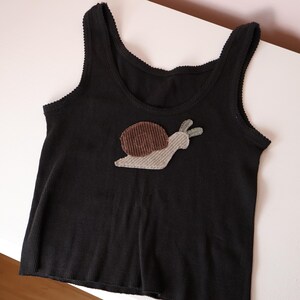 Op de afbeelding: Een zwart geribbelde tanktop met een slak-applicatie op het midden van de borst. De slak heeft een donkerbruin corduroy huisje en een lichtgrijs corduroy lichaam. De tanktop heeft een ronde hals en armsgaten met een fijne schulprand.