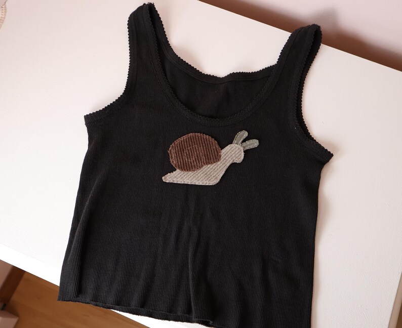 Op de afbeelding: Een zwart geribbelde tanktop met een slak-applicatie op het midden van de borst. De slak heeft een donkerbruin corduroy huisje en een lichtgrijs corduroy lichaam. De tanktop heeft een ronde hals en armsgaten met een fijne schulprand.