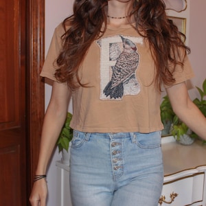 May include: A tan short-sleeved crop top featuring a large rectangular patch on the chest. The patch displays a detailed illustration of a bird perched on a branch, layered over a large letter F. The shirt has a crew neckline and a straight hem.