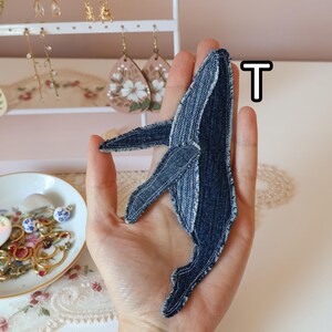 Op de afbeelding: Een walvisvormige stoffen patch gemaakt van donkerblauwe denimstof met zichtbare stiksels langs de randen. De patch heeft een opvallend ontwerp met een vin en staart, waarbij de textuur van de stof goed zichtbaar is. De letter T is in de buurt te zien.