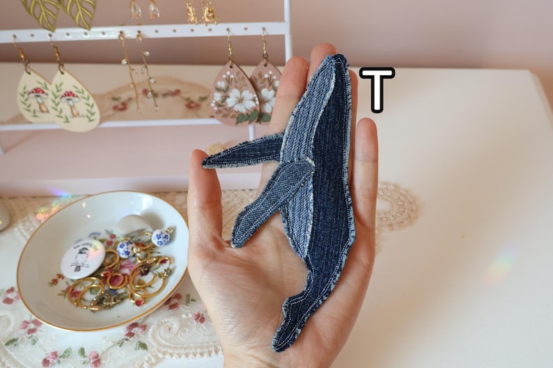 Op de afbeelding: Een walvisvormige stoffen patch gemaakt van donkerblauwe denimstof met zichtbare stiksels langs de randen. De patch heeft een opvallend ontwerp met een vin en staart, waarbij de textuur van de stof goed zichtbaar is. De letter T is in de buurt te zien.