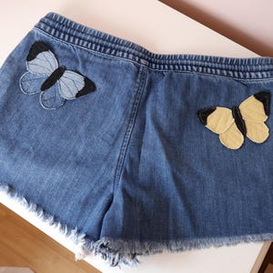 Op de afbeelding: Blauwe spijkershort met een elastische tailleband en een gerafelde zoom. De short is versierd met twee vlindervormige stoffen patches: &eacute;&eacute;n in lichtblauwe en zwarte tinten aan de linkerkant en &eacute;&eacute;n in lichtgele en zwarte tinten aan de rechterkant.