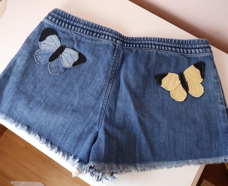 Op de afbeelding: Blauwe spijkershort met een elastische tailleband en een gerafelde zoom. De short is versierd met twee vlindervormige stoffen patches: &eacute;&eacute;n in lichtblauwe en zwarte tinten aan de linkerkant en &eacute;&eacute;n in lichtgele en zwarte tinten aan de rechterkant.