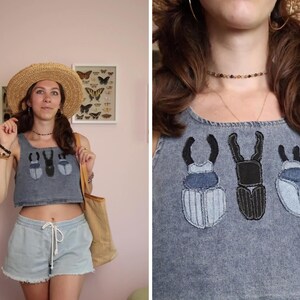 Op de afbeelding: Een grijze acid-wash denim crop top met drie geborduurde kever-patches in verschillende tinten blauw en zwart. De top heeft een vierkante halslijn en wordt gecombineerd met een lichtblauwe denim short met een trekkoord in de taille.
