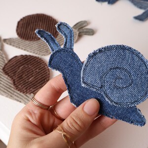 Op de afbeelding: Een slakvormige stoffen patch van blauwe denim met zichtbare gestikte randen en een spiraalvormig schelppatroon. Twee extra slakpatches van bruine en beige corduroy stof zijn zichtbaar op de achtergrond met een vergelijkbaar patroon.