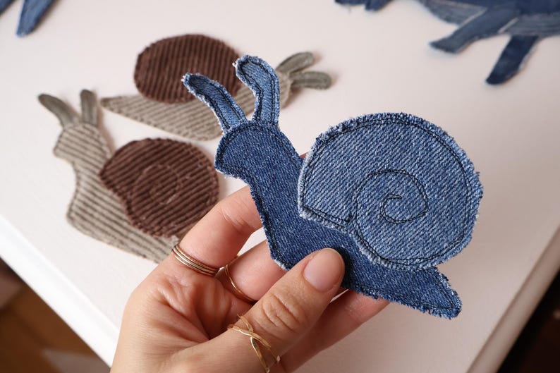 Op de afbeelding: Een slakvormige stoffen patch van blauwe denim met zichtbare gestikte randen en een spiraalvormig schelppatroon. Twee extra slakpatches van bruine en beige corduroy stof zijn zichtbaar op de achtergrond met een vergelijkbaar patroon.