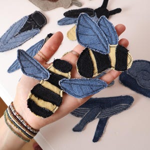 Op de afbeelding: Een verzameling stoffen patches in de vorm van insecten en dieren, waaronder bijen met zwart-geel gestreepte lichamen en blauwe spijkerstof vleugels, een slak, een walvis en andere insectvormen gemaakt van blauwe spijkerstof en zwarte stof.