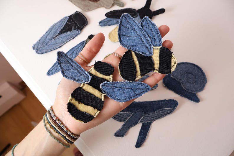 Op de afbeelding: Een verzameling stoffen patches in de vorm van insecten en dieren, waaronder bijen met zwart-geel gestreepte lichamen en blauwe spijkerstof vleugels, een slak, een walvis en andere insectvormen gemaakt van blauwe spijkerstof en zwarte stof.
