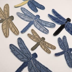 May include: A collection of fabric dragonfly appliques in various shades of blue and tan denim. Each dragonfly features stitched wing details and bodies made from textured fabric, arranged in a scattered pattern across the frame.