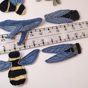 Op de afbeelding: Denim stoffen patches in de vorm van een walvis, twee bijen met gele en zwarte strepen, en twee insecten met blauwe vleugels. Een doorzichtige plastic liniaal ligt over de patches en laat zien dat de walvis ongeveer 11,5 cm lang is.