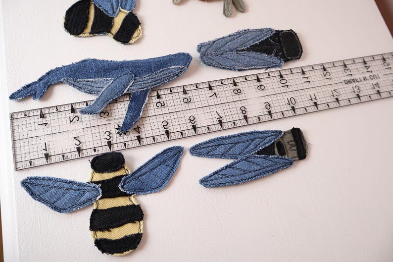 Op de afbeelding: Denim stoffen patches in de vorm van een walvis, twee bijen met gele en zwarte strepen, en twee insecten met blauwe vleugels. Een doorzichtige plastic liniaal ligt over de patches en laat zien dat de walvis ongeveer 11,5 cm lang is.
