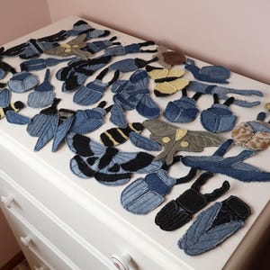 Op de afbeelding: Een verzameling van diverse insectvormige patches gemaakt van blauwe spijkerstof. Het assortiment bevat kevers, motten, vlinders en bijen in verschillende tinten blauw, zwart en beige, voorzien van gestikte details en texturen in de stof.