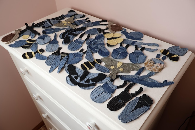 Op de afbeelding: Een verzameling van diverse insectvormige patches gemaakt van blauwe spijkerstof. Het assortiment bevat kevers, motten, vlinders en bijen in verschillende tinten blauw, zwart en beige, voorzien van gestikte details en texturen in de stof.