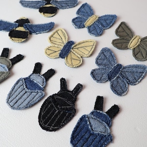 May include: A collection of fabric patches shaped like insects and butterflies, crafted from various shades of denim and cotton. The set includes blue, black, and beige winged creatures with stitched details, frayed edges, and distinct textures.