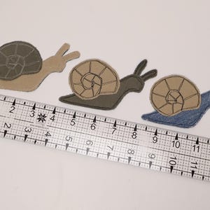 Op de afbeelding: Drie stoffen slak-applicaties met gestikte schelpdetails, geplaatst boven een doorzichtige plastic liniaal met centimeteraanduidingen. De slakken zijn beige, donkergroen en blauw van kleur, elk met een contrasterende kleur voor de slakkenhuisjes.